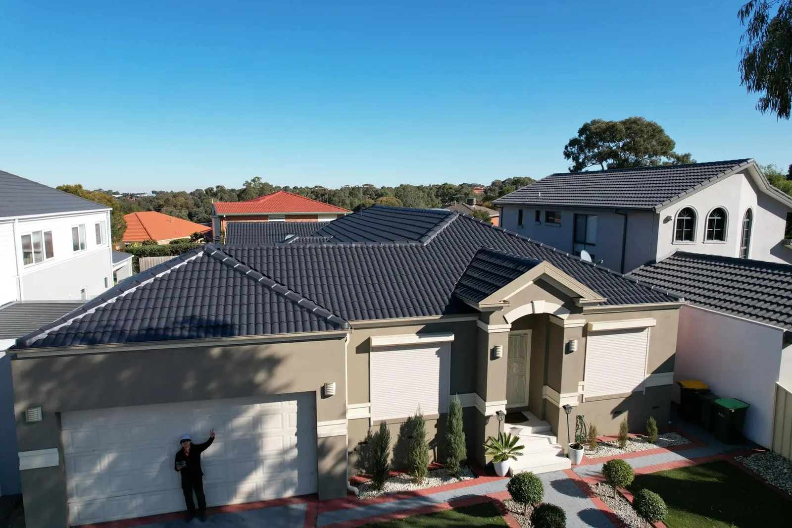 Aerial view of a completed charcoal concrete tile roof restoration on a single-storey brick home, with our roofer waving from the driveway below