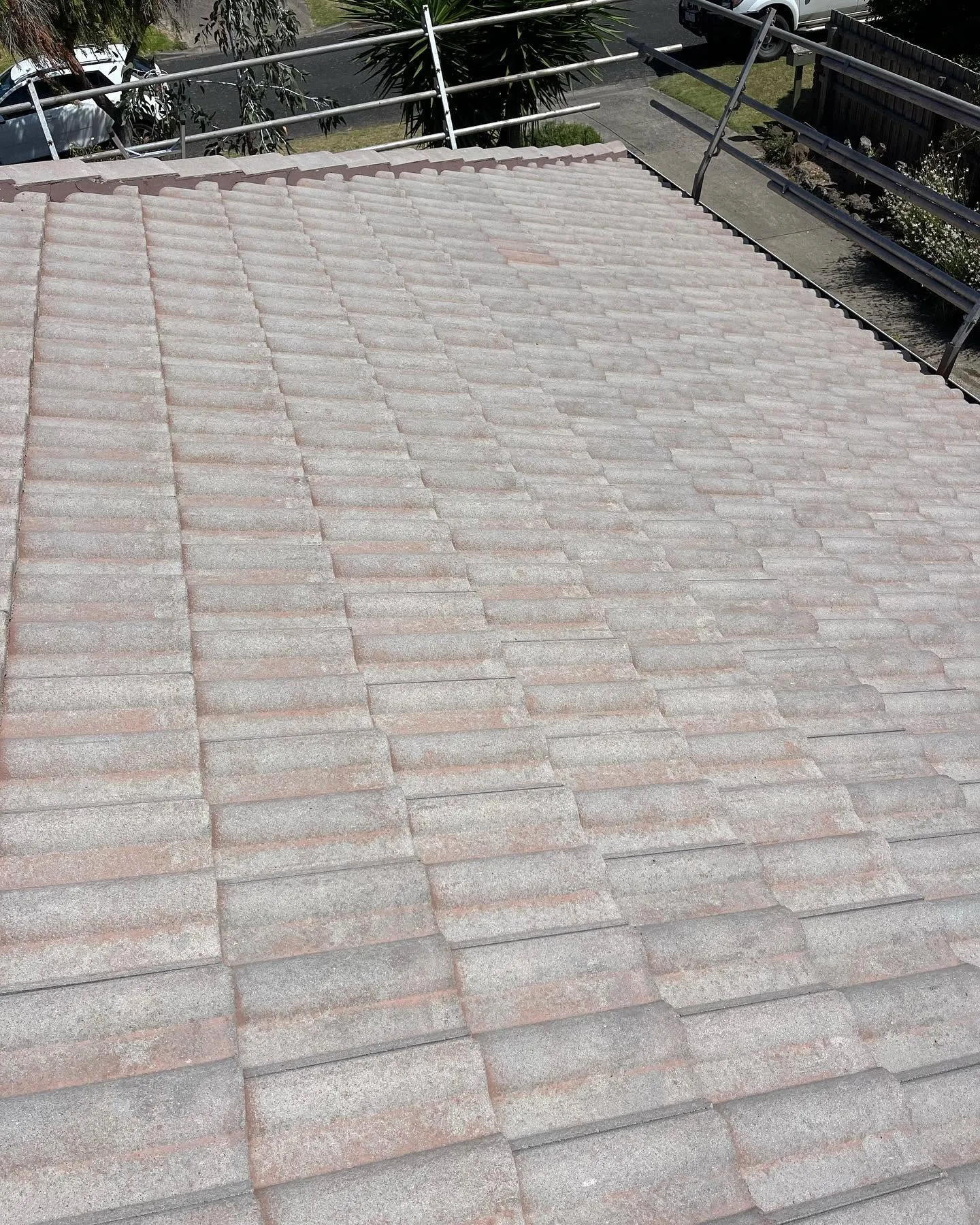 Weathered concrete tile roof viewed from the ridge before cleaning, with scaffolding safety rail and parked vehicles below