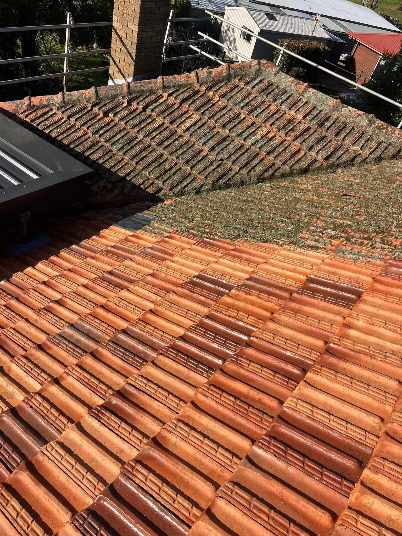Terracotta tile roof mid-clean beside a brick chimney, lichen-covered tiles above and cleaned tiles below
