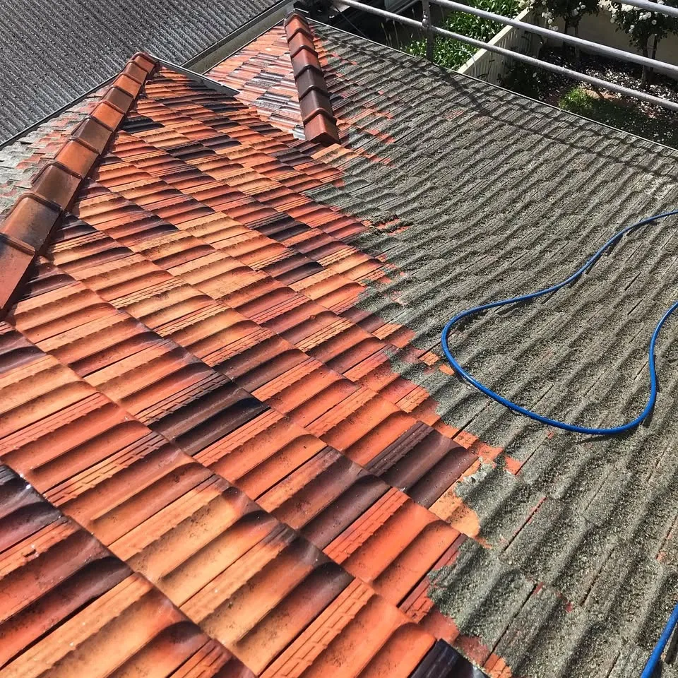 Terracotta tile roof being pressure-washed, hose visible mid-clean between freshly cleaned tiles and lichen-covered tiles