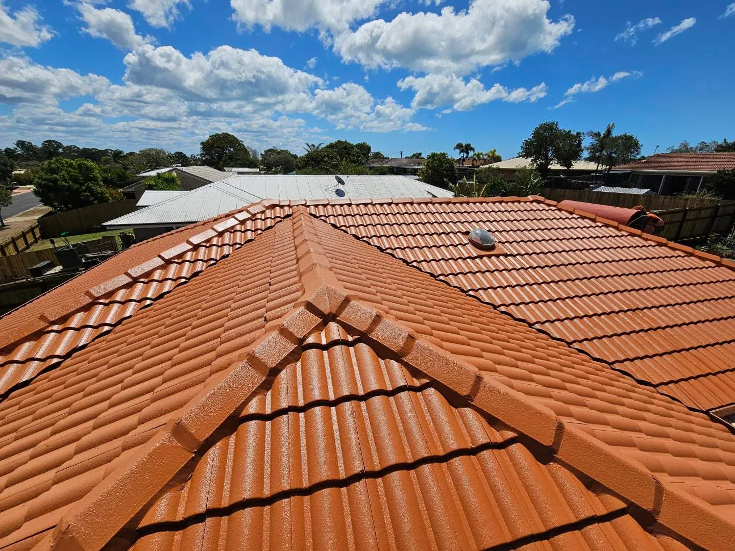 Aerial view of a hip roof freshly painted in dark terracotta, with a roof vent and suburban trees in the background